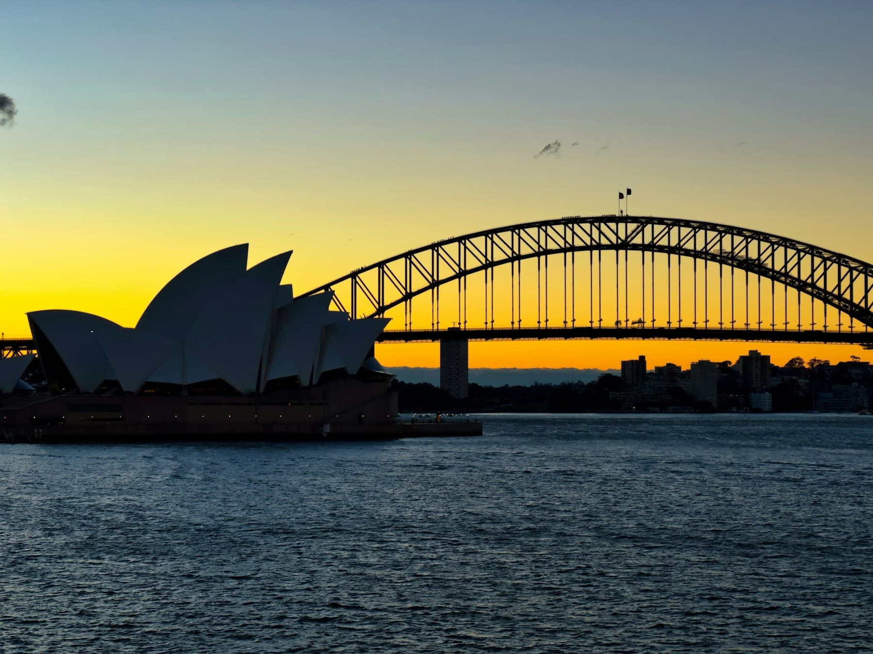 Sydney Harbour at golden hour, Opera House and Harbour Bridge silhouetted against an orange sky.