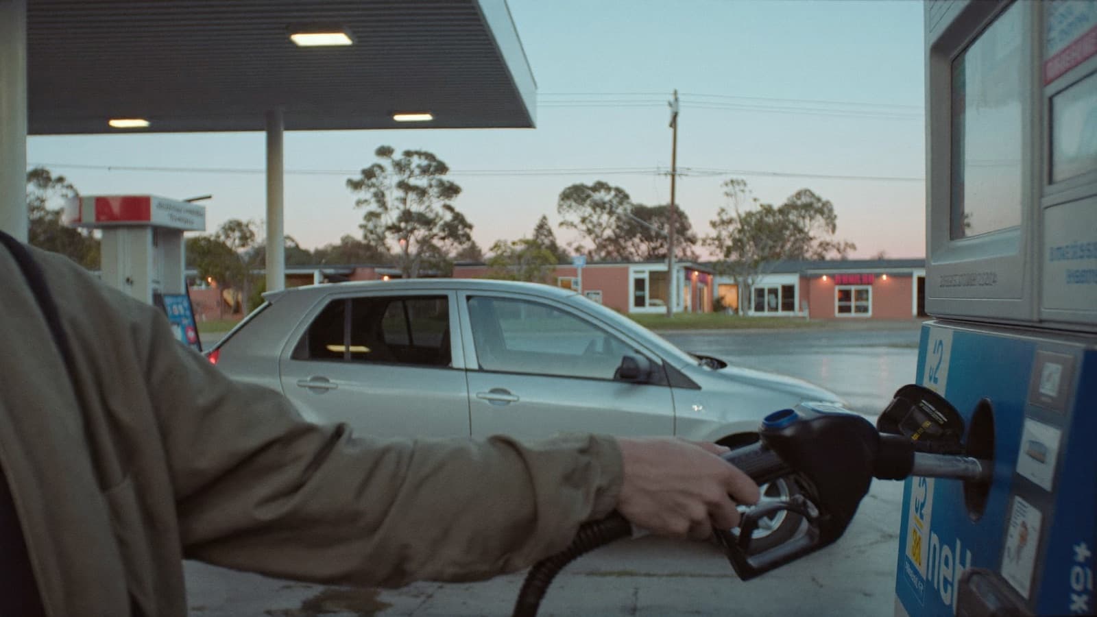 A hand lifting a fuel nozzle at a suburban Australian service station at dawn, with a silver hatchback, brick shops and gum trees behind.