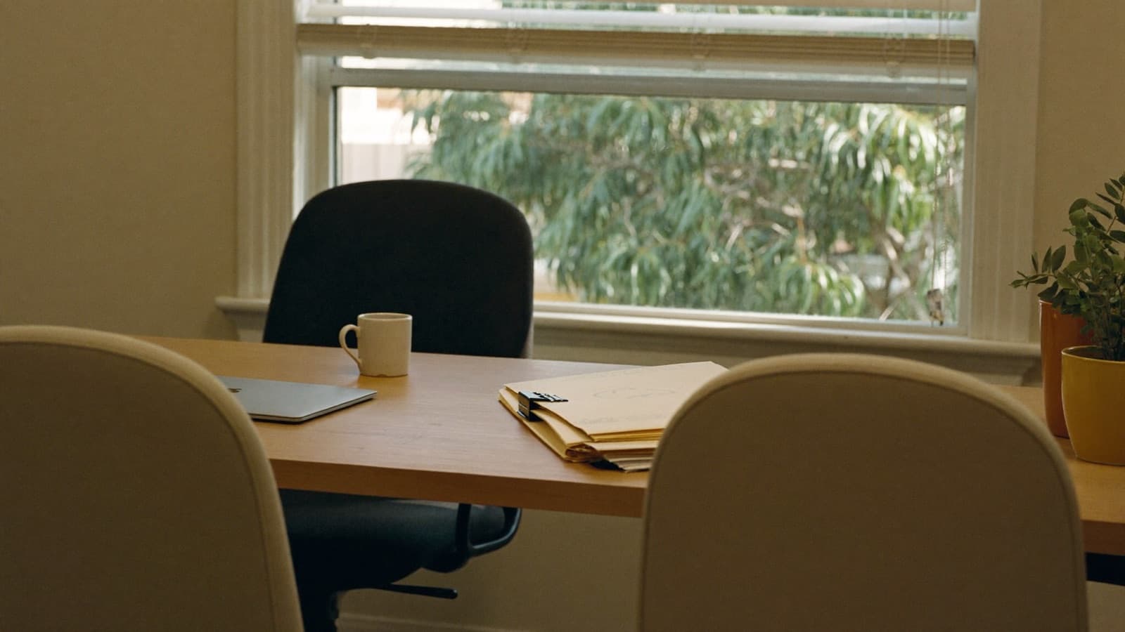 Two empty client chairs facing a broker's desk with a closed laptop, a coffee mug and clipped manila folders, a gum tree outside the office window.