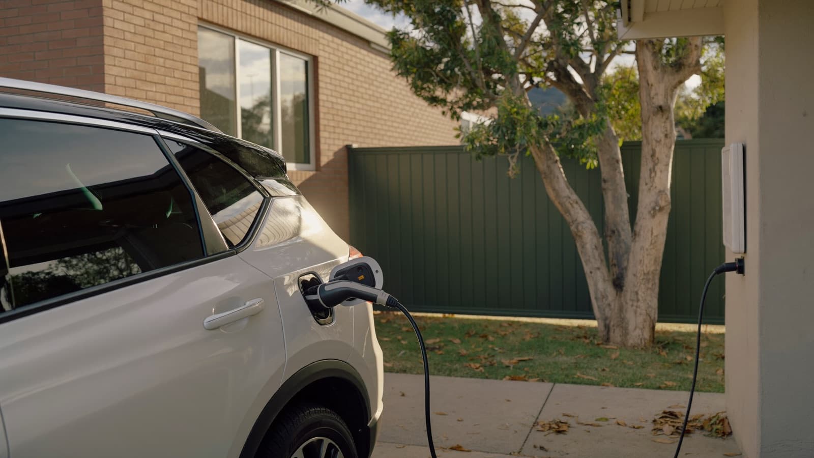 A white electric SUV plugged into a wall charger in the carport of a suburban Australian brick home in late afternoon light.