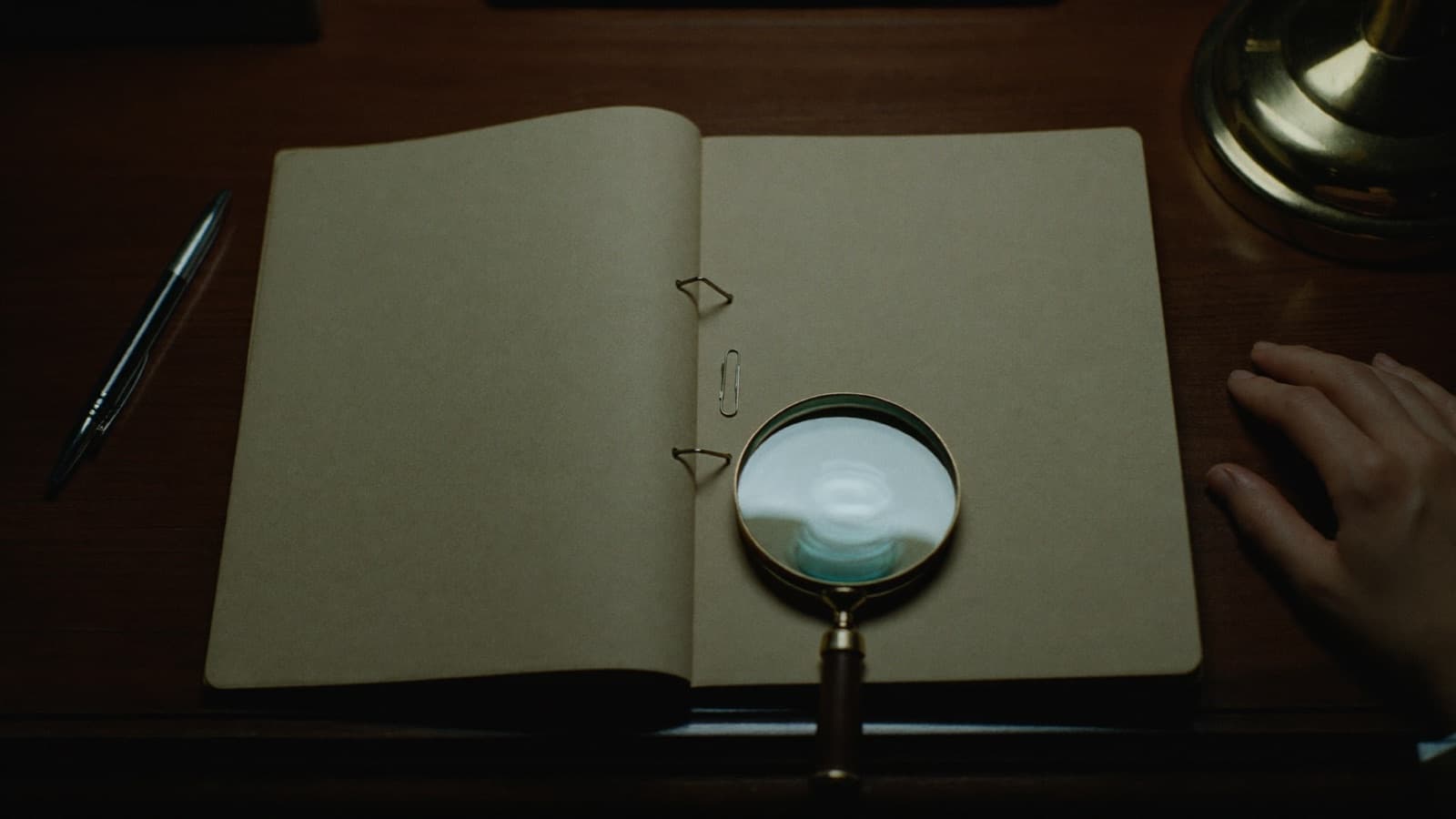 An open manila file under a brass desk lamp at night, with a magnifying glass resting on it and a hand at the edge of the desk.