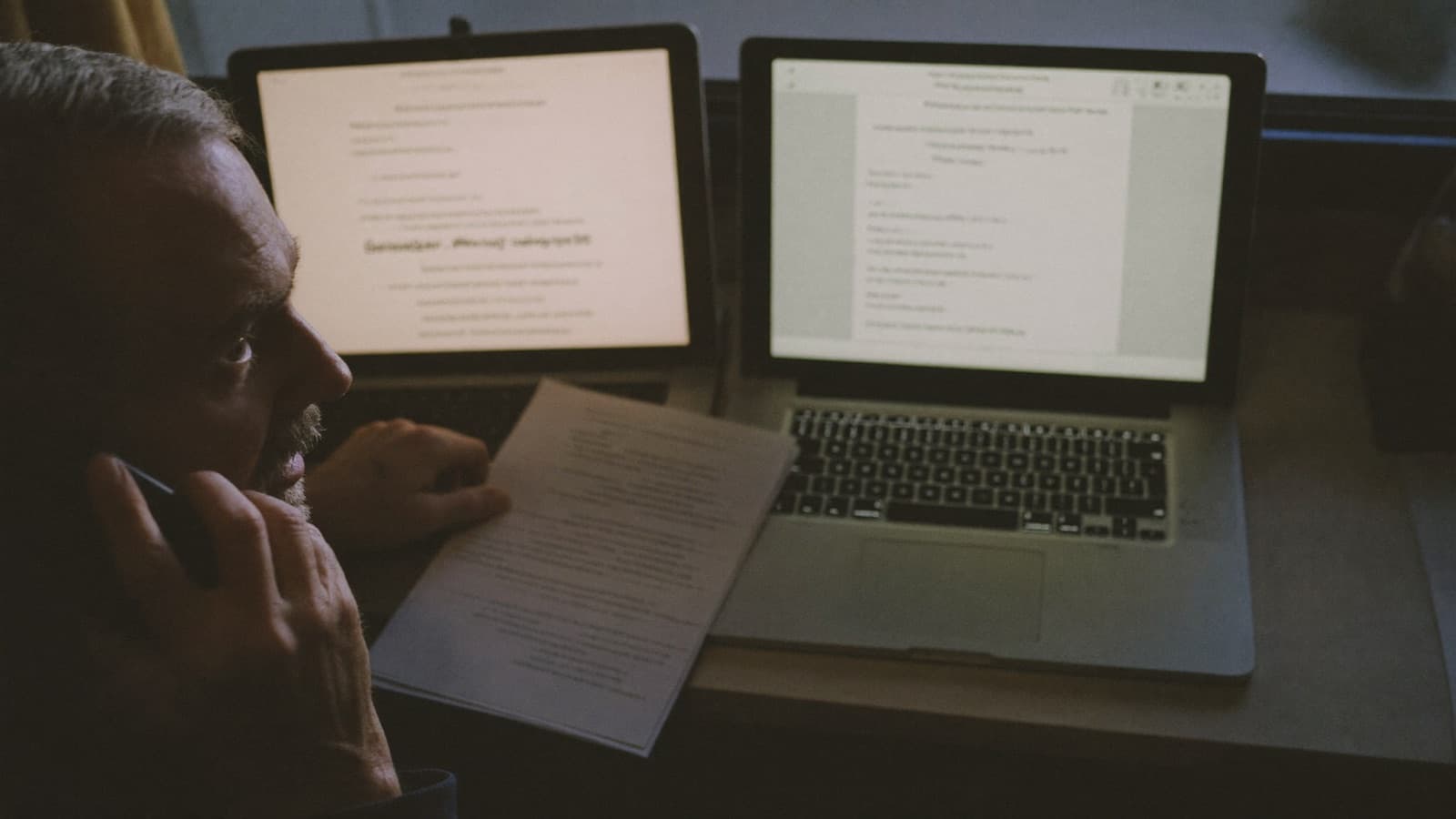 A man at a home office desk at dusk on the phone, comparing two printed statements side by side.