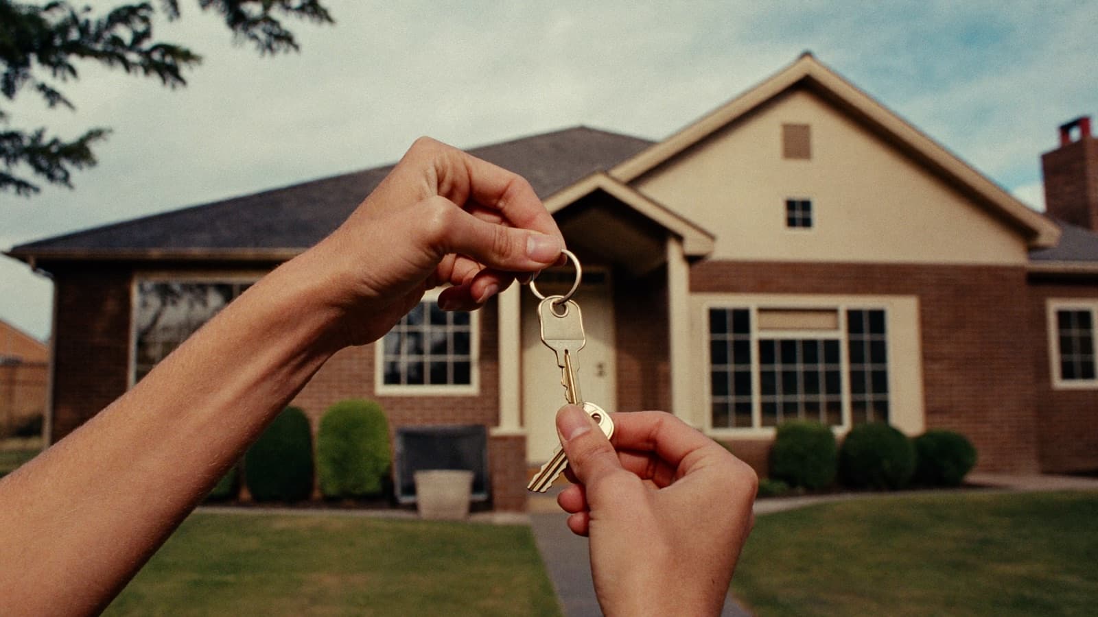 A set of house keys passed from one hand to another over a sold sign on the front lawn of a suburban brick home.