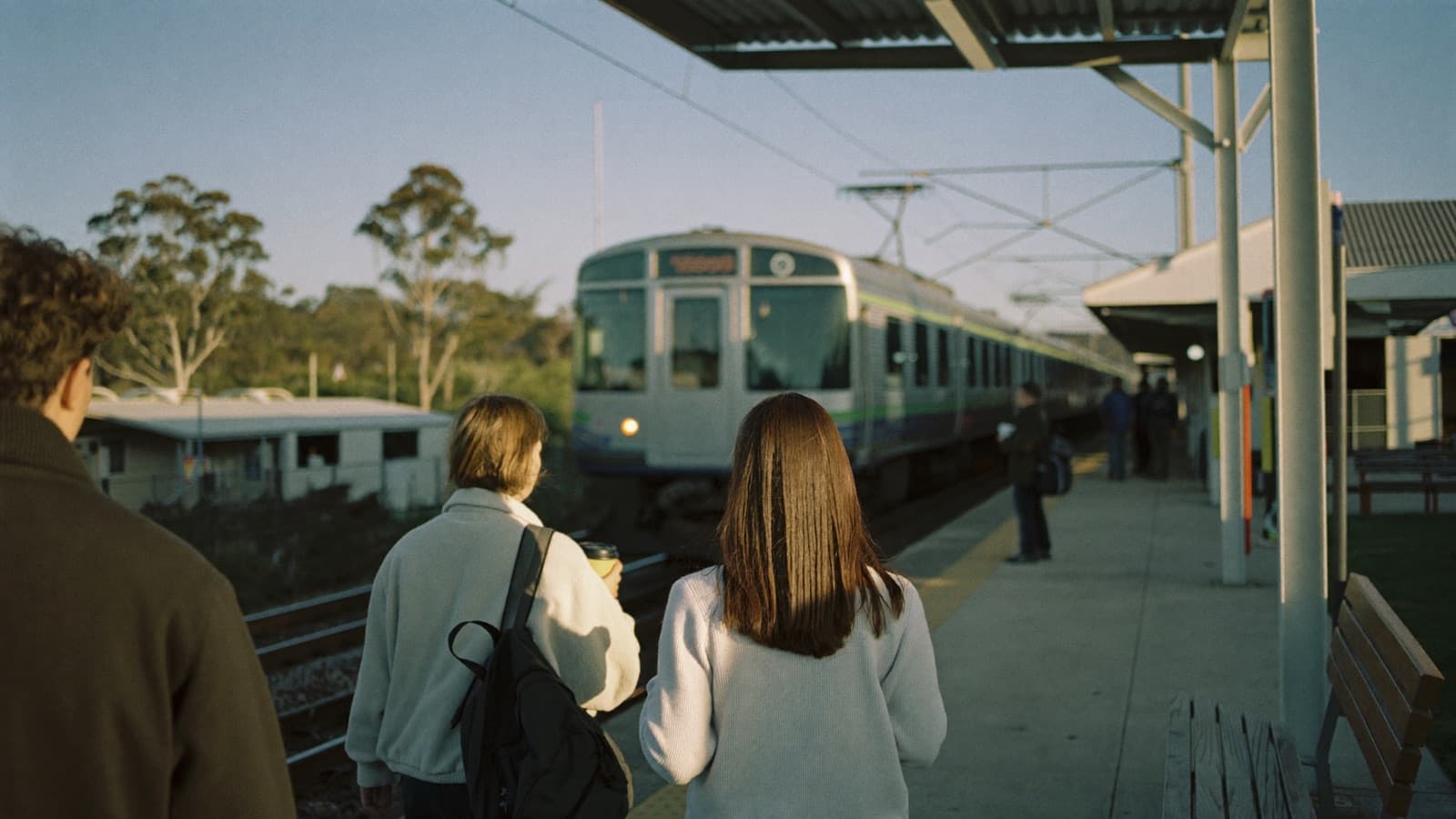 Commuters seen from behind waiting on a suburban Australian train platform in early spring morning light, with a train approaching along the tracks.